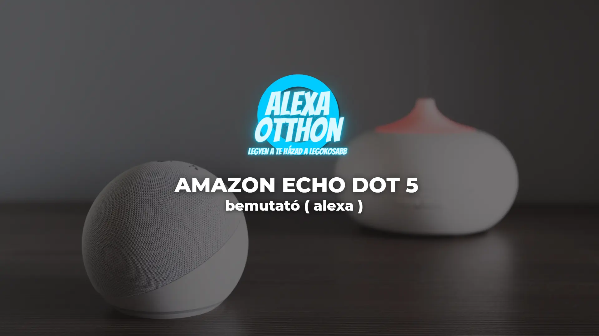 Egy szürke Amazon Echo Dot 5 intelligens hangszóró ül egy sötét felületen, a háttérben egy elmosódott fehér illóolaj diffúzorral. Felettük kék-fehér magyar nyelvű szövegben "ALEXA OTTHON" olvasható, és kiemeli az amazon echo dot 5 bemutatót.